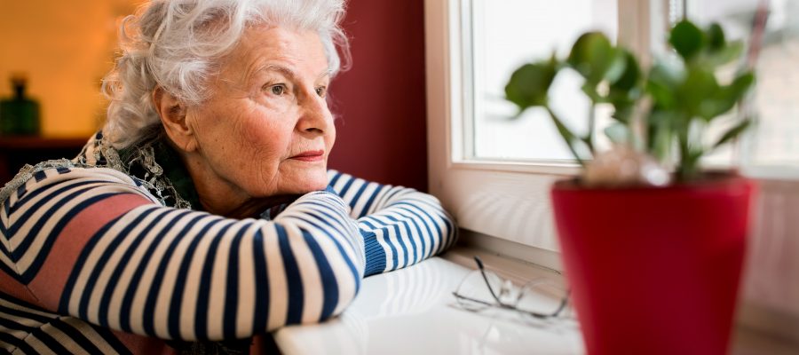 Sad alone senior woman looking through window at home, loneliness concept